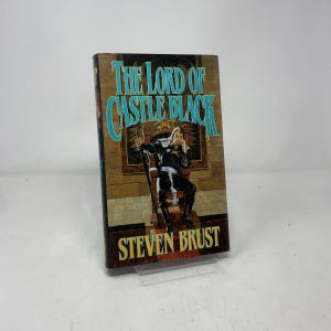 May include: A hardcover book titled "The Lord of Castle Black" by Steven Brust. The cover features a fantasy illustration of a person in black attire seated on a throne. The title is in turquoise letters.