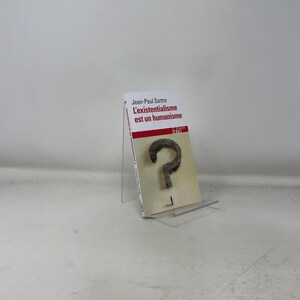 May include: A book titled "L'existentialisme est un humanisme" by Jean-Paul Sartre. The book has a white cover with red accents and a large question mark graphic. The book is displayed on a clear acrylic stand.