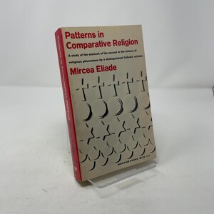 May include: A book titled "Patterns in Comparative Religion" by Mircea Eliade. The book has a beige cover with a repeating pattern of crosses and crescent shapes. The spine is red, and the book is displayed on a clear stand.