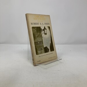 Pode incluir: Um livro vintage intitulado "Chicago Sociology 1920-1932" de Robert E. L. Faris. A capa apresenta uma imagem a preto e branco de um edifício com um candeeiro de rua e o número 1126. O livro é exibido numa base transparente.