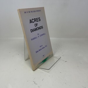 May include: A vintage book titled "Acres of Diamonds" by Russell H. Conwell, displayed on a clear acrylic stand. The book's cover is off-white with black text, including the title and author's name. The book is slightly angled, showcasing its spine and pages.