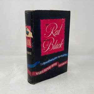 Pode incluir: Um livro preto com um título vermelho e branco, "O Vermelho e o Negro", um romance de Marie-Henri Beyle (Stendhal). A capa do livro apresenta uma fita azul e uma citação sobre a dignidade do amor.