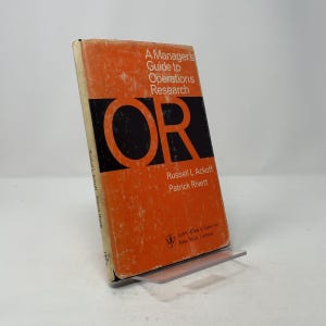 Puede incluir: Un libro antiguo, "A Manager's Guide to Operations Research", con una cubierta naranja y texto negro. El libro se muestra en un soporte transparente, revelando el título y los nombres de los autores. Publicado por John Wiley & Sons.
