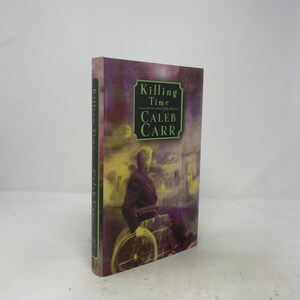 Op de afbeelding: Een hardcover boek getiteld Killing Time van Caleb Carr. De kaft toont een paarse en gele illustratie van een persoon in een rolstoel die naar een stadssilhouet kijkt. De titel en de naam van de auteur staan in het groen op de kaft.