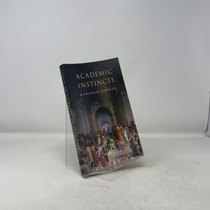 Op de afbeelding: Een boek met de titel "Academic Instincts" van Marjorie Garber. De donkerblauwe kaft heeft gouden letters en een schilderij van mensen in een klassieke setting. Het boek wordt getoond op een heldere acrylstandaard.