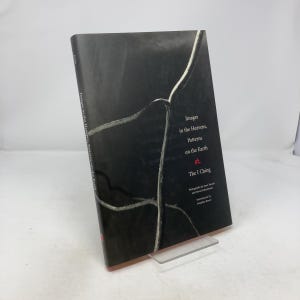 Pode incluir: Um livro com o título "Images in the Heavens, Patterns on the Earth" com capa preta e texto branco e vermelho. O livro está apoiado em um suporte transparente. A lombada do livro é visível.