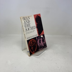 May include: A vintage book titled "Man and the Sacred" by Mircea Eliade. The cover features a black silhouette, red and white accents, and illustrations. The book is propped up on a clear stand.