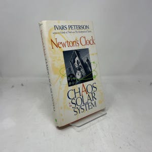 May include: A hardcover book titled "Newton's Clock" by Ivars Peterson. The cover features a black and white image of a clock tower and the title "Chaos in the Solar System". The book is propped up on a clear stand.