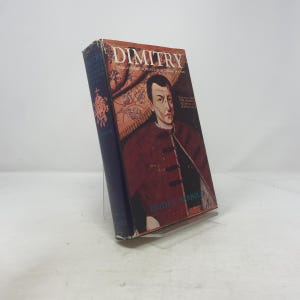 Puede incluir: Un libro de tapa dura titulado "DIMITRY" con un retrato de un hombre con abrigo rojo. El lomo del libro es azul oscuro con letras doradas y un emblema decorativo. El libro está apoyado en un soporte transparente.