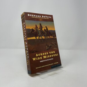 Könnte beinhalten: Ein Hardcover-Buch mit dem Titel "Across the Wide Missouri" von Bernard DeVoto. Das Cover zeigt eine Malerei von Reitern auf Pferden. Das Buch ist ein Gewinner des Pulitzer-Preises.