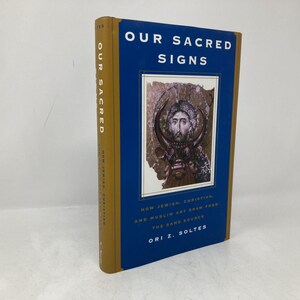 May include: A hardcover book titled "OUR SACRED SIGNS" with a blue cover and gold spine. The book features an image of religious art and text that reads "HOW JEWISH, CHRISTIAN, AND MUSLIM ART DRAW FROM THE SAME SOURCE".