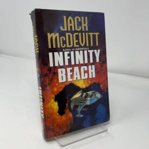 May include: A hardcover book titled Infinity Beach by Jack McDevitt. The cover features a dark blue and orange cosmic background with a small silver spacecraft in the center. The author name is in gold text, and the title is in white block letters.