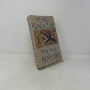 Pode incluir: Um livro de capa dura intitulado "Flying Hero Class" de Thomas Keneally. A capa apresenta uma ilustração estilizada de um avião em preto e vermelho sobre um fundo bege. O livro é exibido em um suporte transparente.