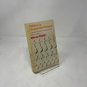 May include: A paperback book titled "Patterns in Comparative Religion" by Mircea Eliade. The cover is beige with orange text and a repeating pattern of black and white shapes. The book is displayed on a clear plastic stand.