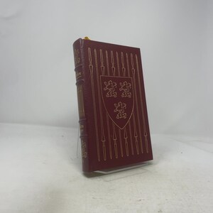 May include: A burgundy-colored book with gold embossed details. The cover features a gold shield with three lions and vertical spear designs. The spine has gold lettering. A yellow ribbon bookmark is visible at the top.