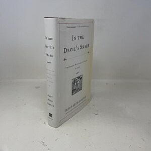 May include: A hardback book titled "In The Devil's Snare" by Mary Beth Norton. The book has a white cover with black text and a small illustration. The book's spine is visible, showing the title and author.