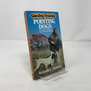 May include: A book titled "Gun-Dog Training Pointing Dogs" with a blue cover featuring a man with a gun and a dog in a field. The book's spine reads "Gun-Dog Training Pointing Dogs" and the author's name is Kenneth C. Roebuck.