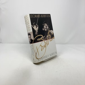 May include: A hardcover book titled "Leonard Bernstein" by Humphrey Burton. The cover features a black and white image of Leonard Bernstein conducting, with his signature in gold. The book is white with black and gold text.