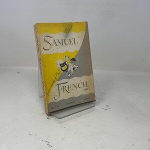 May include: A vintage book titled "Samuel French Inc." with a yellow and gray cover. The cover features the company name, a graphic of theatrical masks, and the text "Basic catalog of Plays!" The book is propped up on a clear stand.