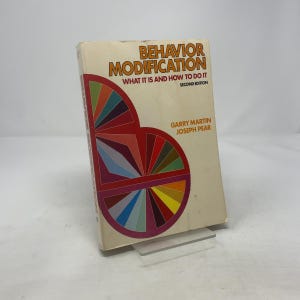 May include: A vintage book titled "Behavior Modification" with the subtitle "What It Is and How to Do It." The cover is cream-colored with orange and red text. A colorful geometric design is on the left side of the book.