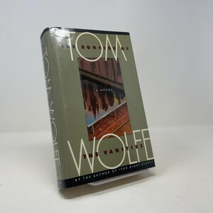 Puede incluir: Un libro de tapa dura titulado The Bonfire of the Vanities de Tom Wolfe. La sobrecubierta es de color verde oliva con letras blancas grandes. Un gráfico rectangular central muestra el reflejo de rascacielos sobre una superficie de cristal y una alfombra.
