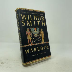 May include: A hardcover book titled Warlock by Wilbur Smith, featuring a black cover with gold lettering and Egyptian-style illustrations of two figures flanking a winged sun disk, with decorative borders at the top and bottom edges.
