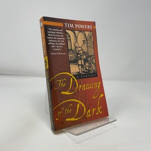 May include: A paperback book titled The Drawing of the Dark by Tim Powers. The cover features a reddish-brown textured background with a black and white illustration of a person in a cellar. The title is written in large, yellow, cursive script.