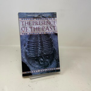 Pode incluir: Um livro intitulado "Morphic Resonance & The Presence of the Past" de Rupert Sheldrake. A capa apresenta uma imagem de fóssil em tons de cinza e azul, com texto branco e vermelho. O livro é exibido em um suporte transparente.