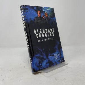 May include: A hardcover book titled Standard Candles by Jack McDevitt. The cover features a dark blue space-themed illustration with two figures, a large satellite dish, and a small silhouette of a person standing against a starry background.