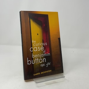 以下が含まれることがあります： ガブリエル・ブラウンスタイン著『The Curious Case of Benjamin Button apt. 3W』のペーパーバック。表紙は、暗い茶色の枠に囲まれた鮮やかな黄色のドアが特徴で、小さな黒い覗き穴があり、暖かく薄暗い背景が描かれています。