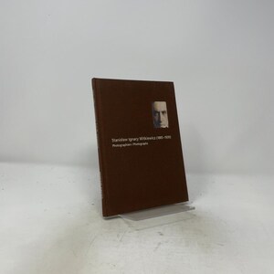 May include: A brown hardcover book titled Stanisław Ignacy Witkiewicz (1885-1939) Photographien / Photographs. The front cover features a small, rectangular black and white portrait of a man centered near the top right corner of the book.