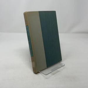 Puede incluir: Un libro antiguo con una cubierta turquesa y beige, exhibido en un soporte acrílico transparente. La columna vertebral presenta texto en un rectángulo turquesa. Las páginas del libro muestran desgaste.