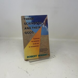 Op de afbeelding: Een hardcover boek getiteld "Three Scientists and Their Gods" van Robert Wright. De omslag heeft tekst in een vetgedrukte letter, met een blauwe lucht en een oranje driehoekontwerp. Het boek wordt op een transparante standaard weergegeven.