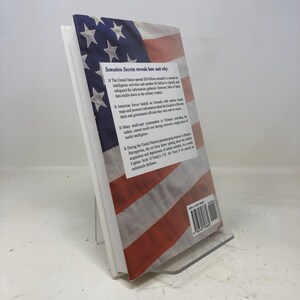 May include: A hardcover book titled "Senseless Secrets" with a cover design featuring the American flag. The book is propped up on a clear display stand, showcasing the title and a summary of the book's content.