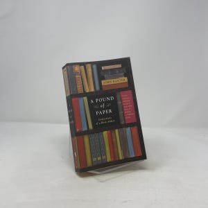May include: The book "A Pound of Paper: Confessions of a Book Addict" is shown, with a cover designed to look like a bookshelf. The spines of the books are in various colours. The author's name, John Baxter, is also visible on the cover.