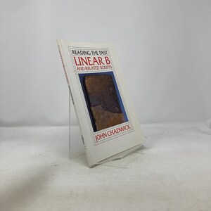 May include: A white book titled "Reading the Past Linear B and Related Scripts" by John Chadwick. The cover features a brown image of a tablet. The book is displayed on a clear acrylic stand.