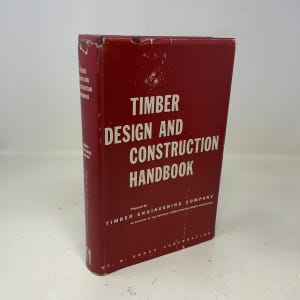 May include: A red hardcover book titled "TIMBER DESIGN AND CONSTRUCTION HANDBOOK" with white lettering. The book is slightly worn, with visible wear on the edges. The book is prepared by Timber Engineering Company.