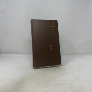 Op de afbeelding: Een bruin hardcover boek met de titel "The Nature and Properties of Soils" van Nyle C. Brady. De titel en auteur van het boek zijn in het geel op de voorkant gedrukt. Het boek staat rechtop op een doorzichtige plastic standaard.