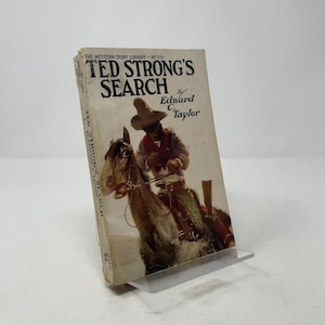 Może przedstawiać: Książka w miękkiej oprawie w stylu vintage zatytułowana "Ted Strong's Search" autorstwa Edwarda C. Taylora. Okładka przedstawia ilustrację w sepii przedstawiającą kowboja na koniu. Część "The Western Story Library - No 25".