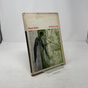 以下が含まれることがあります： Naum Gaboの「Of Divers Arts」というタイトルのヴィンテージハードカバー本。表紙には、ローブを着た人物の白黒イラストが描かれており、緑と白のアクセントが施されています。本は透明なスタンドに展示されています。