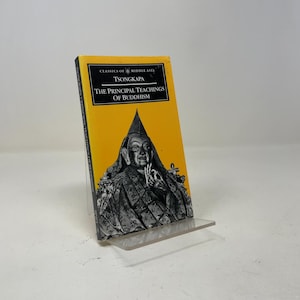 May include: A paperback book titled The Principal Teachings of Buddhism by Tsongkapa, part of the Classics of Middle Asia series. The cover features a bright yellow background with a black and white illustration of a figure in traditional robes.