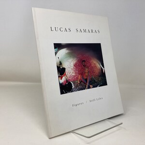 May include: A white book cover with the title "Lucas Samaras Figures / Still Lifes". The cover features a photograph of a man in a room with pink and white walls.