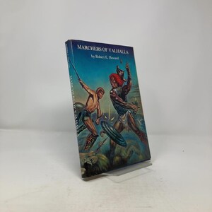 Könnte beinhalten: Ein Vintage-Hardcover-Buch mit dem Titel "Marchers of Valhalla" von Robert E. Howard. Das Cover zeigt eine Illustration von zwei Kriegern im Kampf, vor blauem Himmel und einer Landschaft mit einem Löwen.