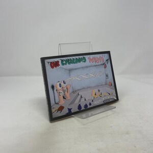May include: A framed artwork with the text "OUR EYEBEAMS TWISTE 7". The artwork features a surreal illustration with cartoon eyes, a wavy beam, and abstract figures in a room. The frame is black, and the artwork is displayed on a clear stand.