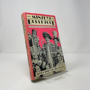Puede incluir: Un libro antiguo titulado "Minsky's Burlesque" con una portada rosa. La portada presenta una imagen en blanco y negro de artistas y el texto "A Fast and Funny Look at America's Bawdiest Era". El libro está apoyado en un soporte transparente.