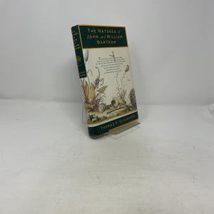 May include: A hardcover book titled "The Natures of John and William Bartram" with a green and gold cover. The cover features illustrations of plants and aquatic life. The author's name, Thomas P. Slaughter, is at the bottom.
