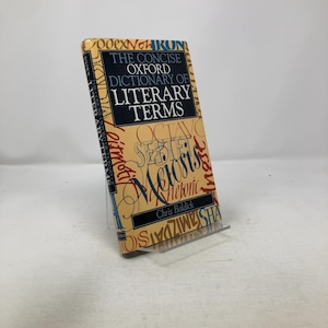 Peut inclure: Un livre de poche intitulé "The Concise Oxford Dictionary of Literary Terms" de Chris Baldick. La couverture est beige avec du texte noir et bleu et des motifs décoratifs. Le livre est présenté sur un présentoir en plastique transparent.