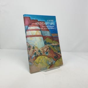 May include: A book titled "A More Abundant Life: New Deal Artists and Public Art in New Mexico" by Jacqueline Hoefer. The cover features a colorful painting of a landscape with red, orange, yellow, and green hills and a blue sky.