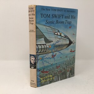 May include: A vintage paperback book cover featuring a blue and white illustration of a futuristic spaceship flying over a city. The book title is "Tom Swift and His Sonic Boom Trap" and the author is Victor Appleton II.