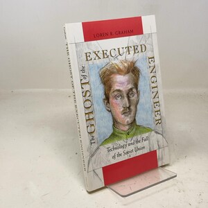 May include: A hardcover book titled "Executed Engineer" by Loren R. Graham. The cover features a portrait of a man and the text "The Ghost of the Executed Engineer". The book is propped up on a clear plastic stand.